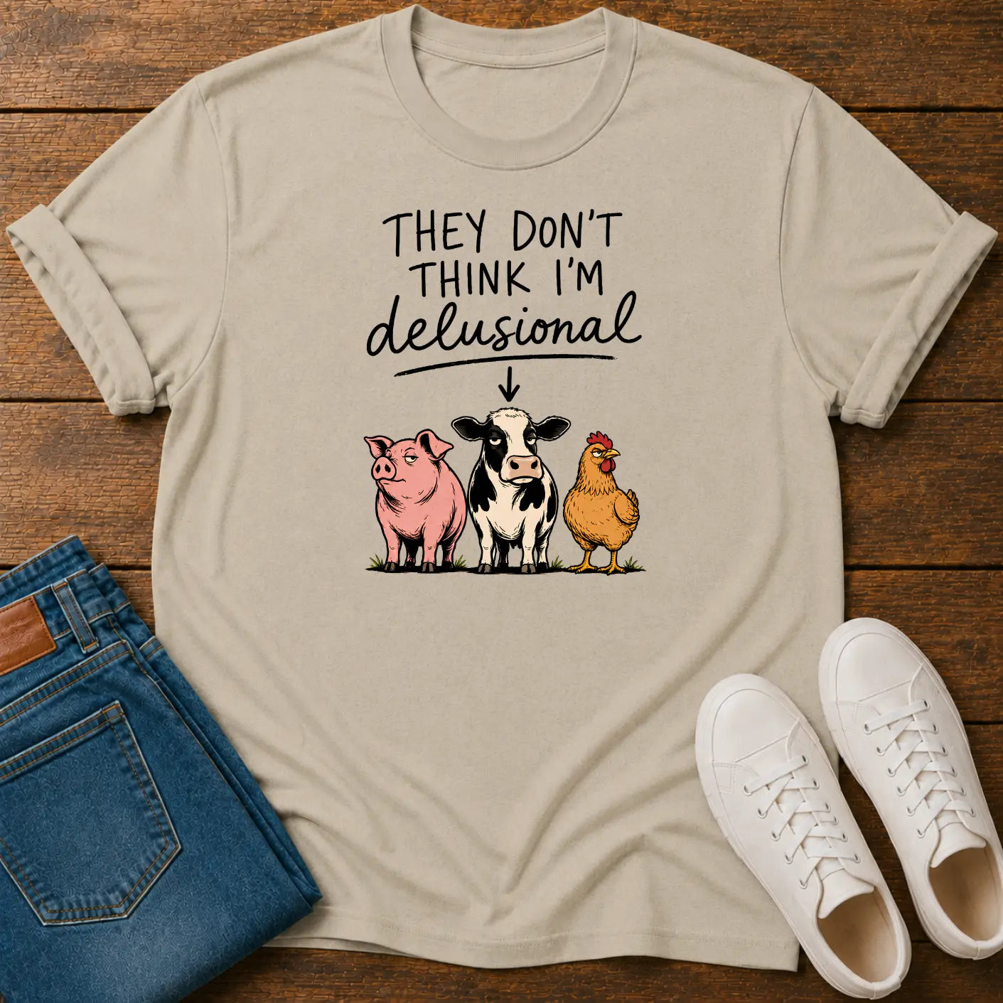 A beige graphic t-shirt laid flat on a wooden surface, styled with folded blue jeans and a pair of white sneakers. The t-shirt features a cartoon graphic of a pink pig, a black-and-white cow, and a yellow chicken under the text 'THEY DON'T THINK I'M delusional'.