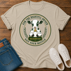 A beige graphic t-shirt laid flat on a wooden surface, styled with folded blue jeans and white sneakers. The t-shirt features a circular graphic of a black-and-white cow with a bell and the text 'FOR THE RIGHTFUL OWNER RESERVED NOT YOUR BOTTLE'.
