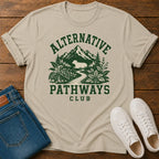 A beige short-sleeve graphic T-shirt laid flat on a wooden surface, styled with folded blue jeans and white sneakers. The shirt has a dark green outdoor-style print reading “ALTERNATIVE PATHWAYS CLUB” with mountains, trees, and a winding path.