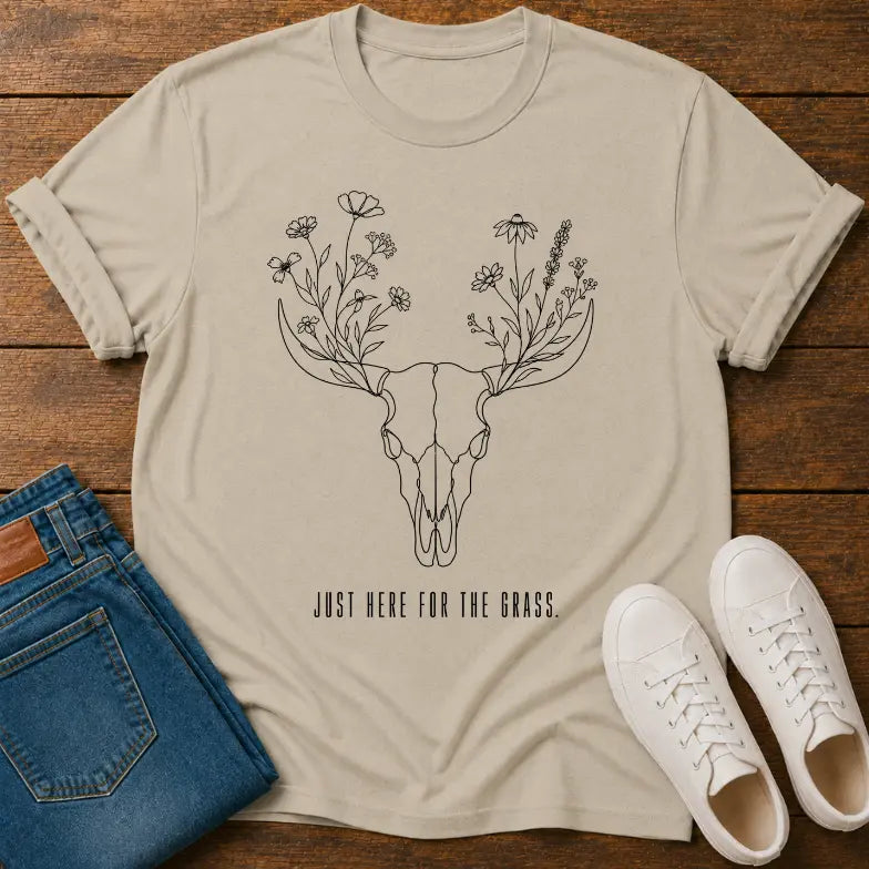 A beige graphic t-shirt laid flat on a wooden surface, styled with folded blue jeans and white sneakers. The t-shirt features a black line art design of a deer skull with flowers growing from its antlers and the text 'JUST HERE FOR THE GRASS.