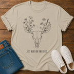 A beige graphic t-shirt laid flat on a wooden surface, styled with folded blue jeans and white sneakers. The t-shirt features a black line art design of a deer skull with flowers growing from its antlers and the text 'JUST HERE FOR THE GRASS.