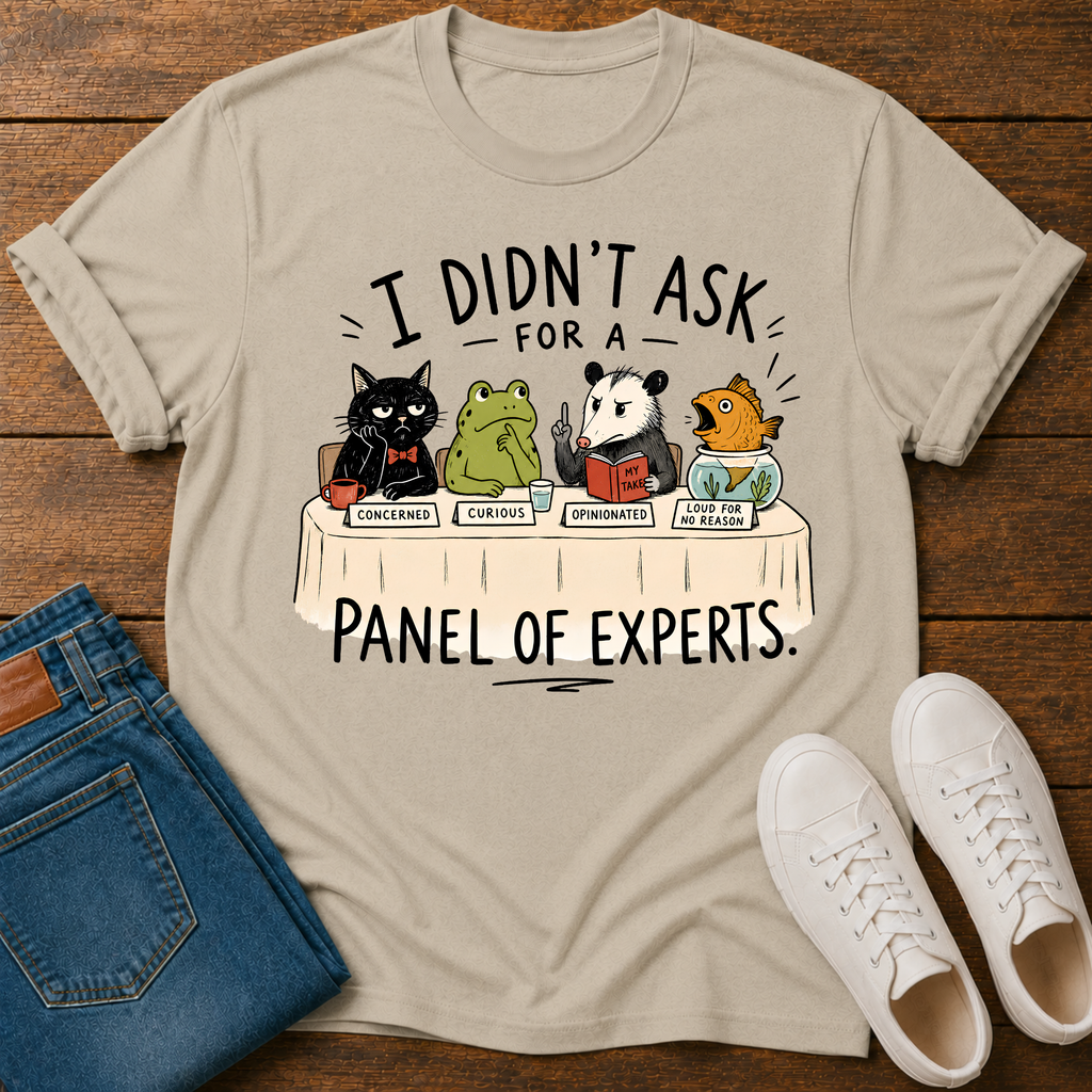A beige graphic t-shirt laid flat on a wooden surface, styled with folded blue jeans and white sneakers. The t-shirt features a cartoon illustration of five animals (a black cat, green frog, panda, goldfish, and orange fish) sitting at a table labeled 'PANEL OF EXPERTS' with humorous responses to 'I DIDN'T ASK FOR A' PANEL OF EXPERTS'