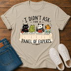 A beige graphic t-shirt laid flat on a wooden surface, styled with folded blue jeans and white sneakers. The t-shirt features a cartoon illustration of five animals (a black cat, green frog, panda, goldfish, and orange fish) sitting at a table labeled 'PANEL OF EXPERTS' with humorous responses to 'I DIDN'T ASK FOR A' PANEL OF EXPERTS'