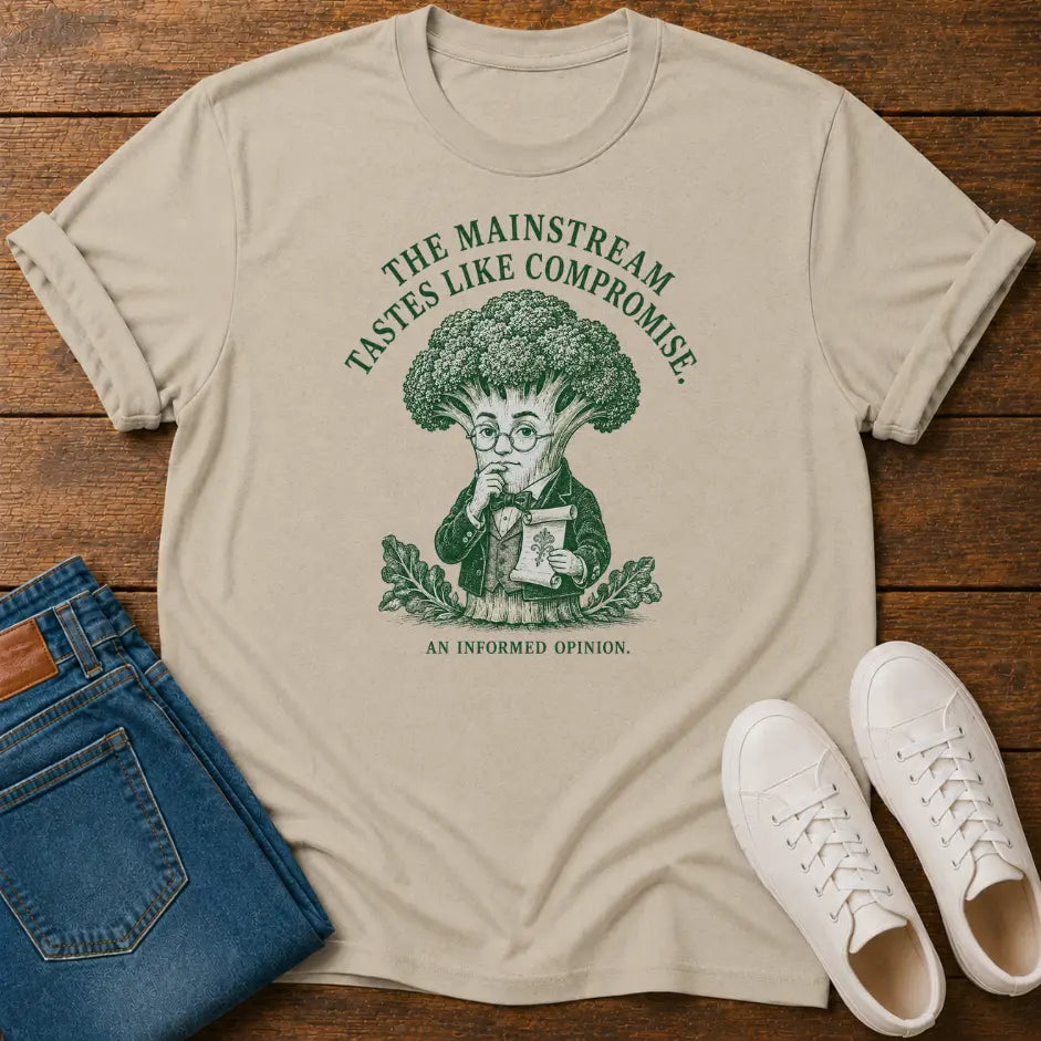 A beige graphic t-shirt featuring a green illustration of a broccoli-headed character and the text 'THE MAINSTREAM TASTES LIKE COMPROMISE. AN INFORMED OPINION.