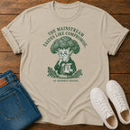 A beige graphic t-shirt featuring a green illustration of a broccoli-headed character and the text 'THE MAINSTREAM TASTES LIKE COMPROMISE. AN INFORMED OPINION.