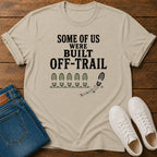 A beige graphic t-shirt laid flat on a wooden surface, styled with blue jeans and white sneakers. The t-shirt features the black text 'SOME OF US WERE BUILT OFF-TRAIL' above a graphic of bootprints.
