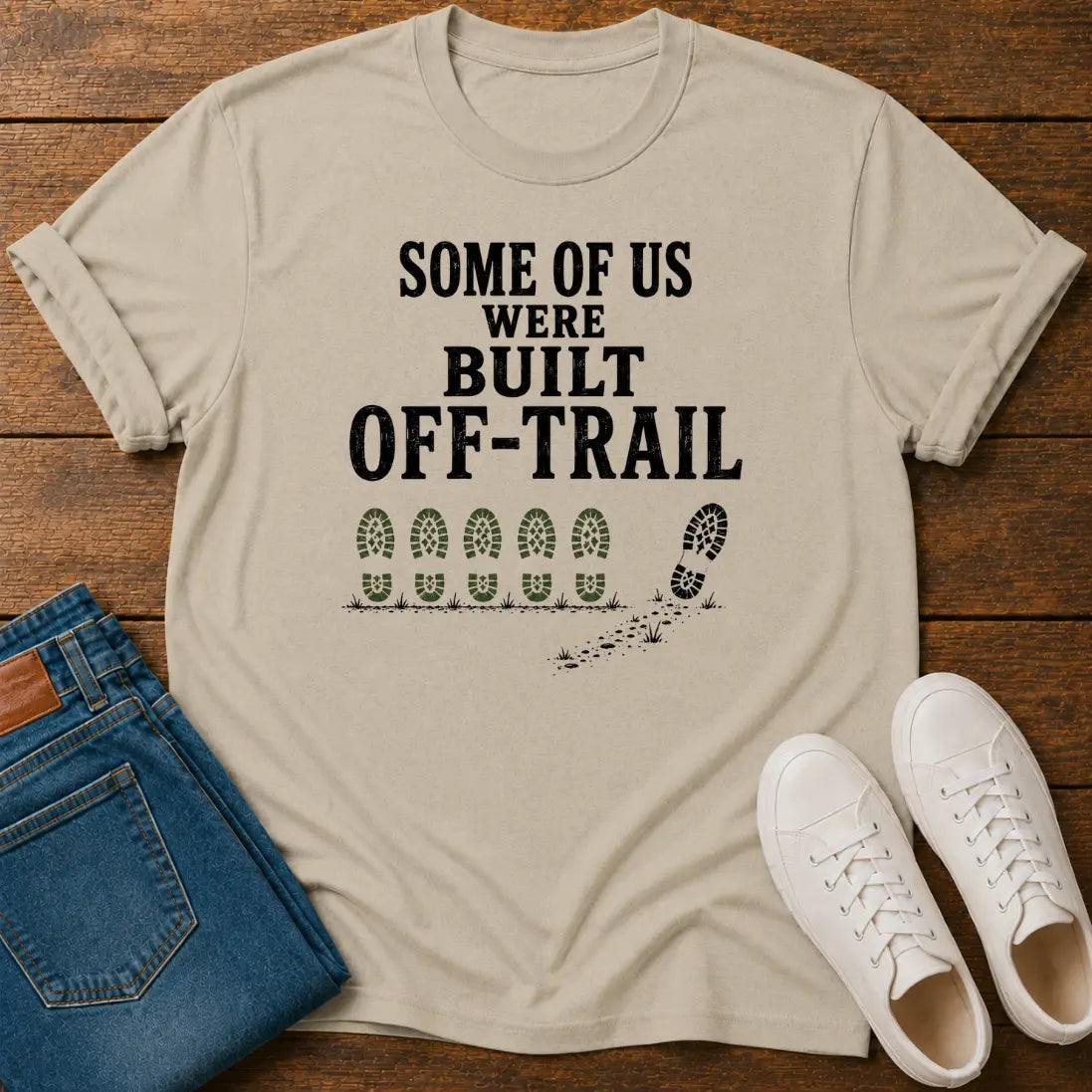 A beige graphic t-shirt laid flat on a wooden surface, styled with blue jeans and white sneakers. The t-shirt features the black text 'SOME OF US WERE BUILT OFF-TRAIL' above a graphic of bootprints.