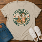 A sand colored graphic t-shirt laid flat on a wooden surface, styled with folded blue jeans and a pair of white sneakers. The t-shirt features a circular retro graphic with the text 'SURVIVED THE HYPOTHETICAL ISLAND' and an illustration of a woman and a pig on a beach.
