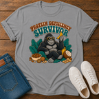 A light gray graphic t-shirt laid flat on a wooden surface, styled with blue jeans and white sneakers. The t-shirt features a retro-style illustration of a gorilla wearing sunglasses and holding a banana, surrounded by leaves and food items, with the text 'PROTEIN DEFICIENCY SURVIVOR' above it.