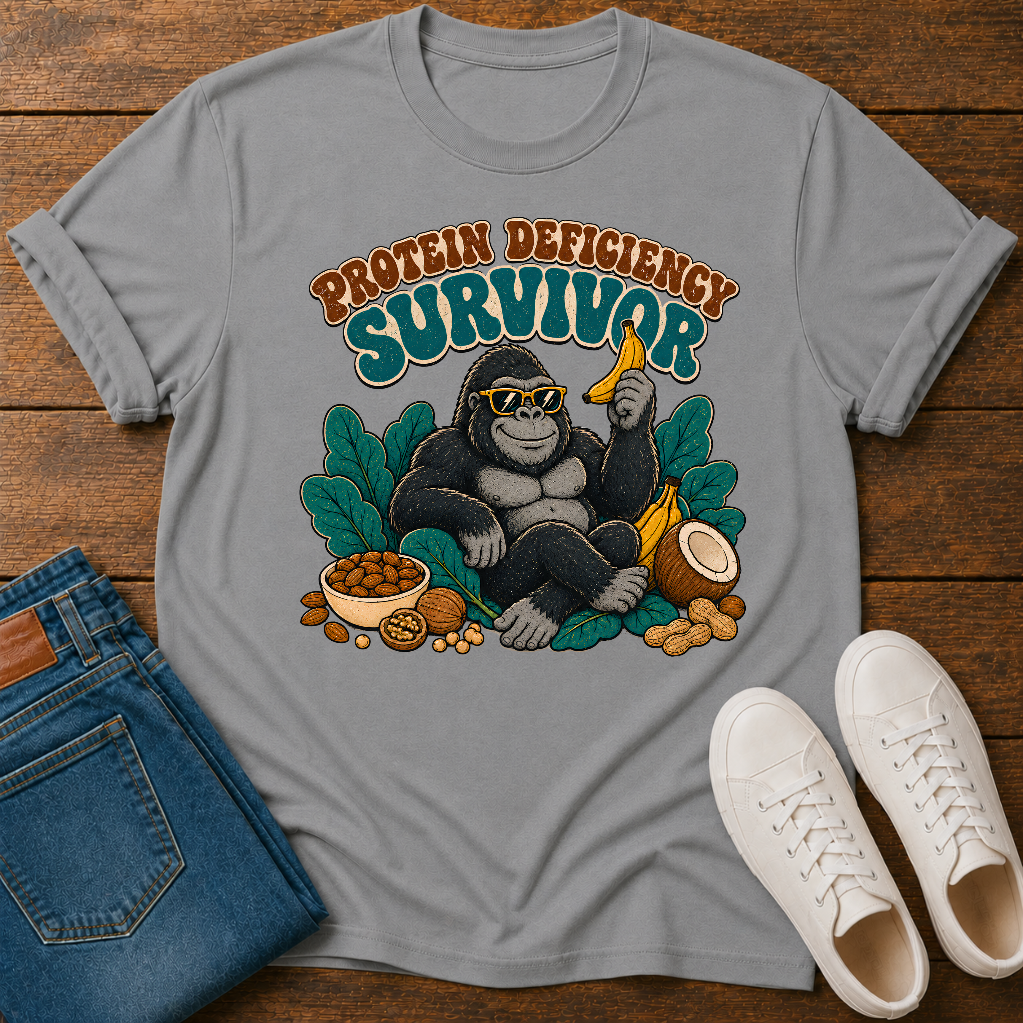 A light gray graphic t-shirt laid flat on a wooden surface, styled with blue jeans and white sneakers. The t-shirt features a retro-style illustration of a gorilla wearing sunglasses and holding a banana, surrounded by leaves and food items, with the text 'PROTEIN DEFICIENCY SURVIVOR' above it.