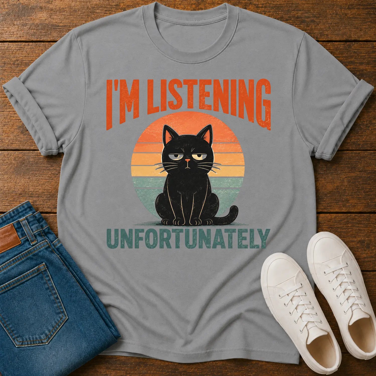 A gray short-sleeve graphic T-shirt laid flat on a wooden surface, styled with folded blue jeans and white sneakers. The shirt has a retro sunset-style cat graphic with the text “I'M LISTENING UNFORTUNATELY” in orange and teal.