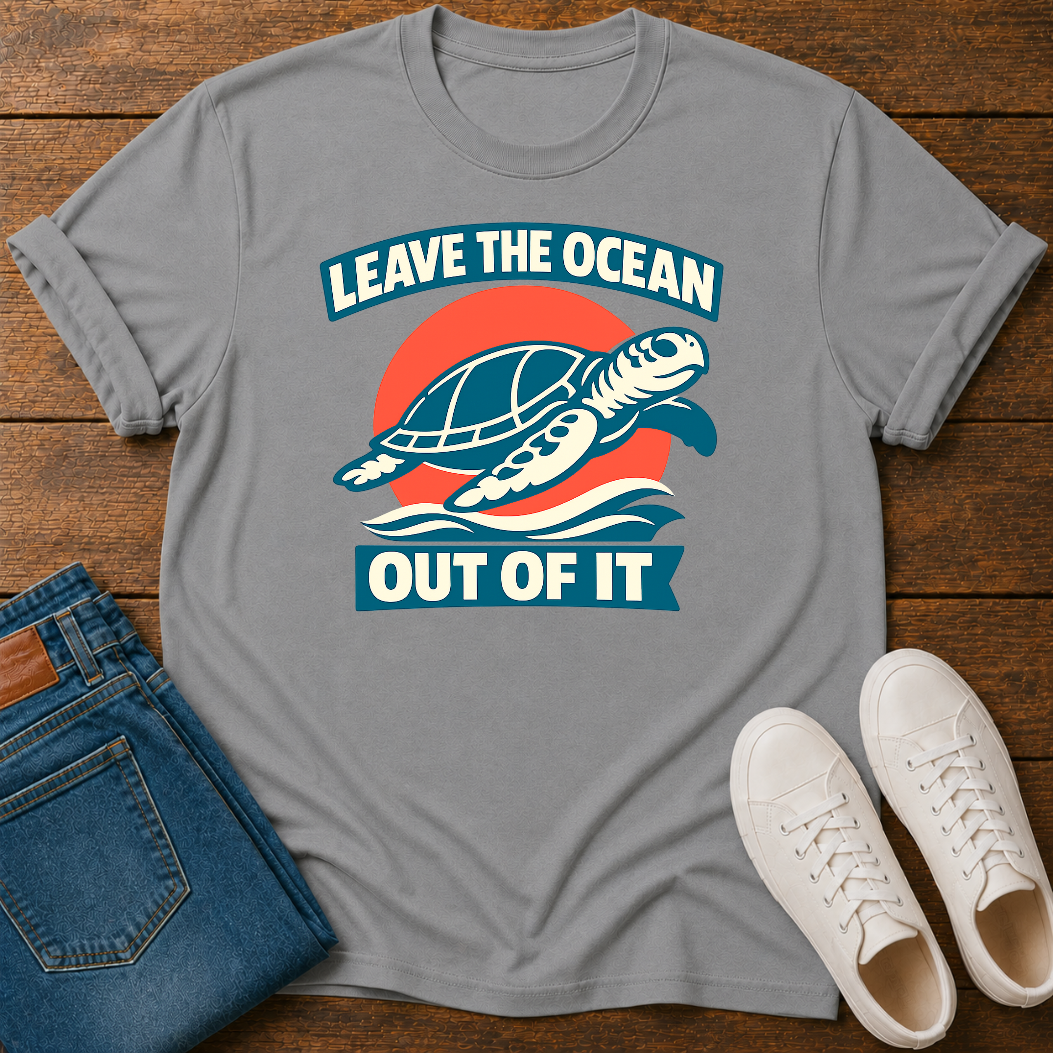 A gray short-sleeve graphic T-shirt laid flat on a wooden surface, styled with folded blue jeans and a pair of white sneakers. The shirt features a turtle illustration with the text 'LEAVE THE OCEAN OUT OF IT' in a retro blue, red, and white design.