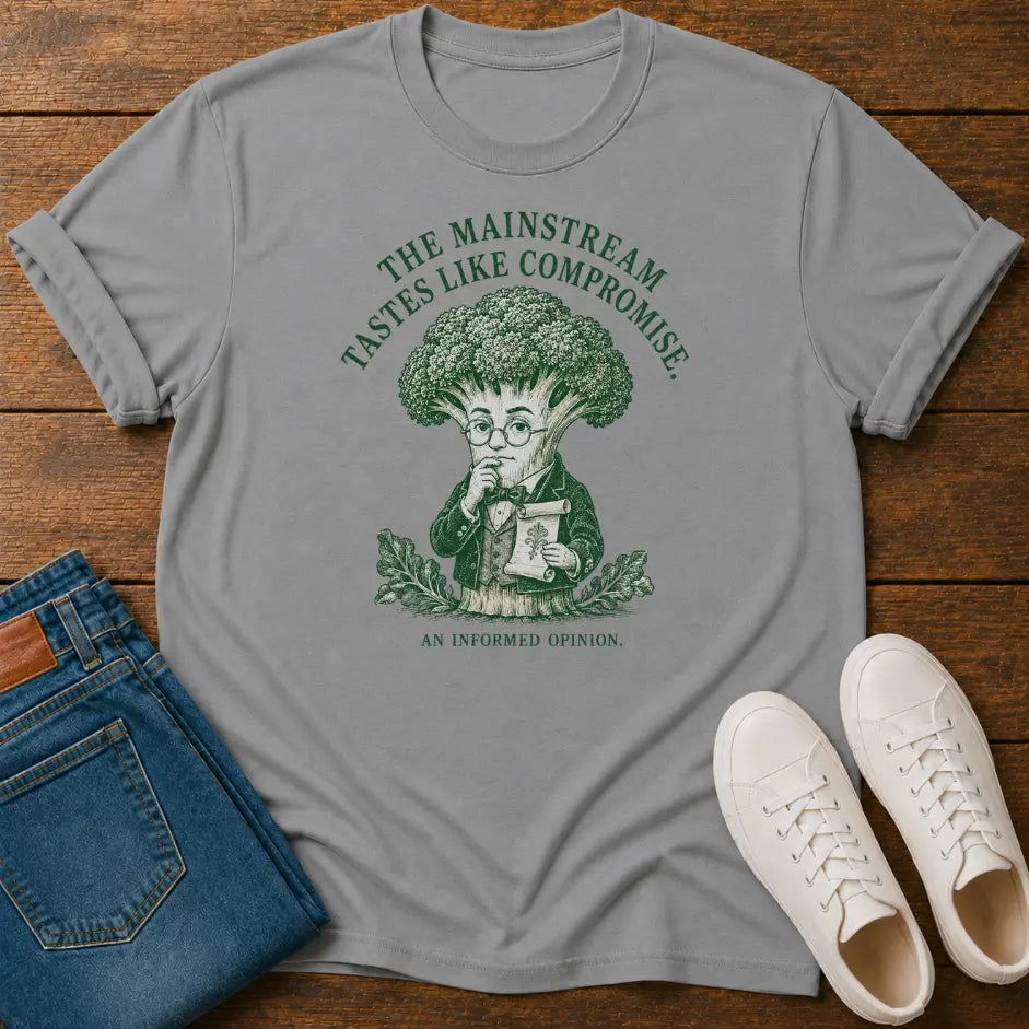 A light gray graphic t-shirt featuring a green illustration of a broccoli character and the text 'THE MAINSTREAM TASTES LIKE COMPROMISE. AN INFORMED OPINION.