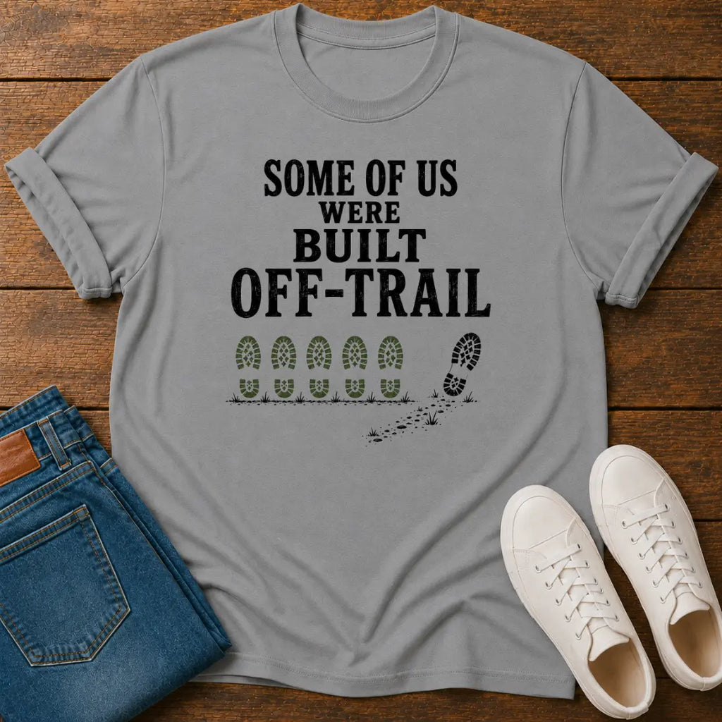A light gray graphic t-shirt laid flat on a wooden surface, styled with folded blue jeans and a pair of white sneakers. The t-shirt features the black text 'SOME OF US WERE BUILT OFF-TRAIL' above a row of green and black footprints.