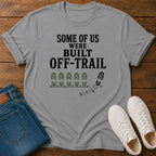 A light gray graphic t-shirt laid flat on a wooden surface, styled with folded blue jeans and a pair of white sneakers. The t-shirt features the black text 'SOME OF US WERE BUILT OFF-TRAIL' above a row of green and black footprints.