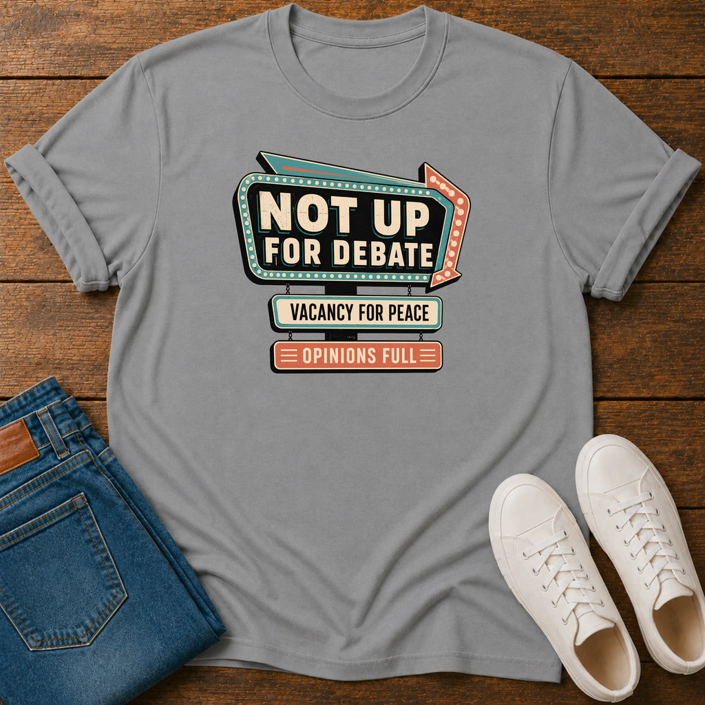 A gray short-sleeve graphic T-shirt laid flat on a wooden surface, styled with folded blue jeans and a pair of white sneakers. The shirt features a retro marquee-style graphic reading 'NOT UP FOR DEBATE' with additional text below.