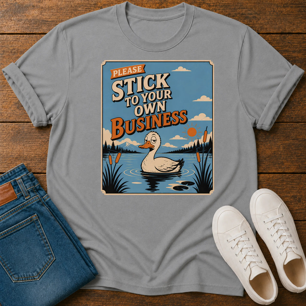 A light gray crewneck t-shirt featuring a retro-style graphic of a duck in a pond with the text 'PLEASE STICK TO YOUR OWN BUSINESS'. The shirt is styled in a flat-lay arrangement on a wooden surface alongside blue jeans and white sneakers.