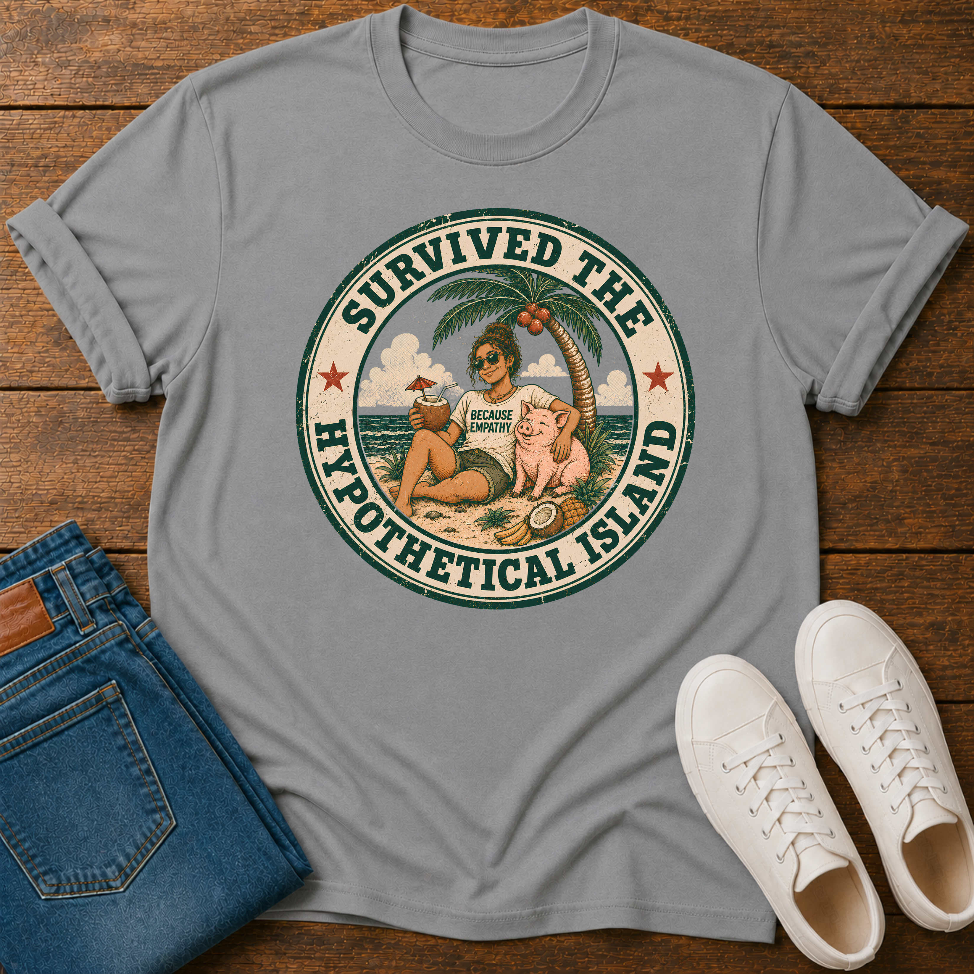 A light gray crewneck t-shirt with a circular retro graphic reading 'SURVIVED THE HYPOTHETICAL ISLAND' featuring a woman and pig on a beach. The shirt is styled in a flat lay on a wooden surface alongside blue jeans and white sneakers.