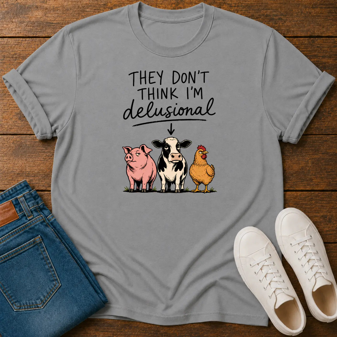 A light gray graphic t-shirt laid flat on a wooden surface, styled with folded blue jeans and white sneakers. The t-shirt features a humorous farm-themed graphic with the text 'THEY DON'T THINK I'M delusional' above an illustration of a pink pig, a black-and-white cow, and a yellow chicken.
