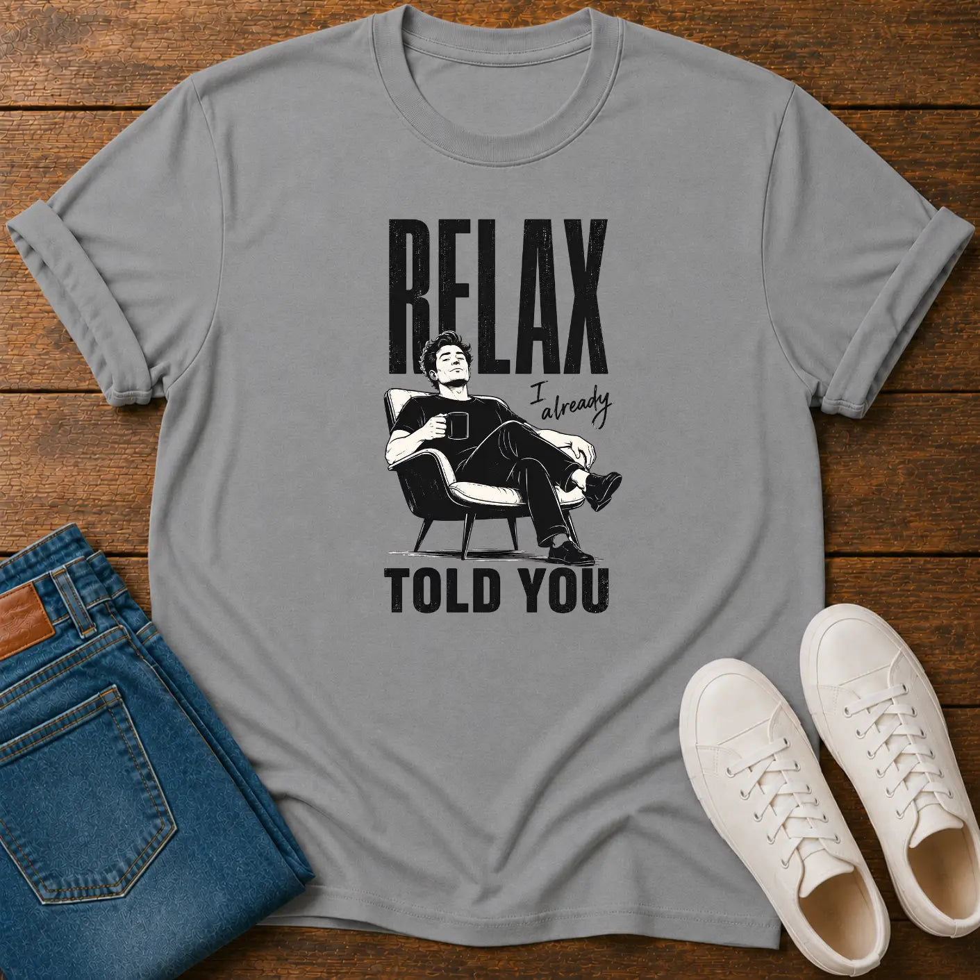 A gray short-sleeve graphic T-shirt laid flat on a wooden surface, styled with folded blue jeans and white sneakers. The shirt has a black printed design featuring the text 'RELAX Already TOLD YOU' and an illustration of a man sitting in a chair.