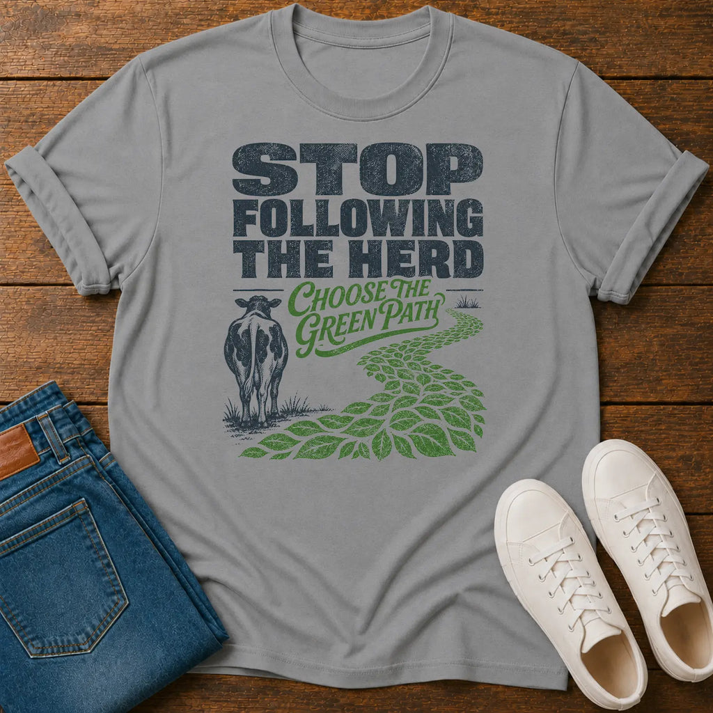 A light gray graphic t-shirt laid flat on a wooden surface, styled with folded blue jeans and white sneakers. The t-shirt features a graphic with the text 'STOP FOLLOWING THE HERD' and 'CHOOSE THE GREEN PATH' alongside a cow and a green leafy path.