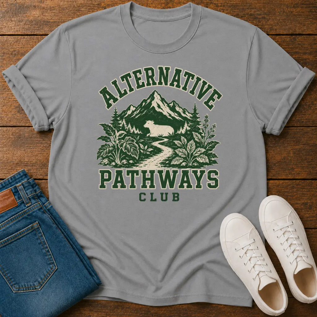 A light gray short-sleeve t-shirt with a green and cream graphic reading 'ALTERNATIVE PATHWAYS CLUB' over a mountain and forest scene, styled flat-lay on a wooden surface with blue jeans and white sneakers.