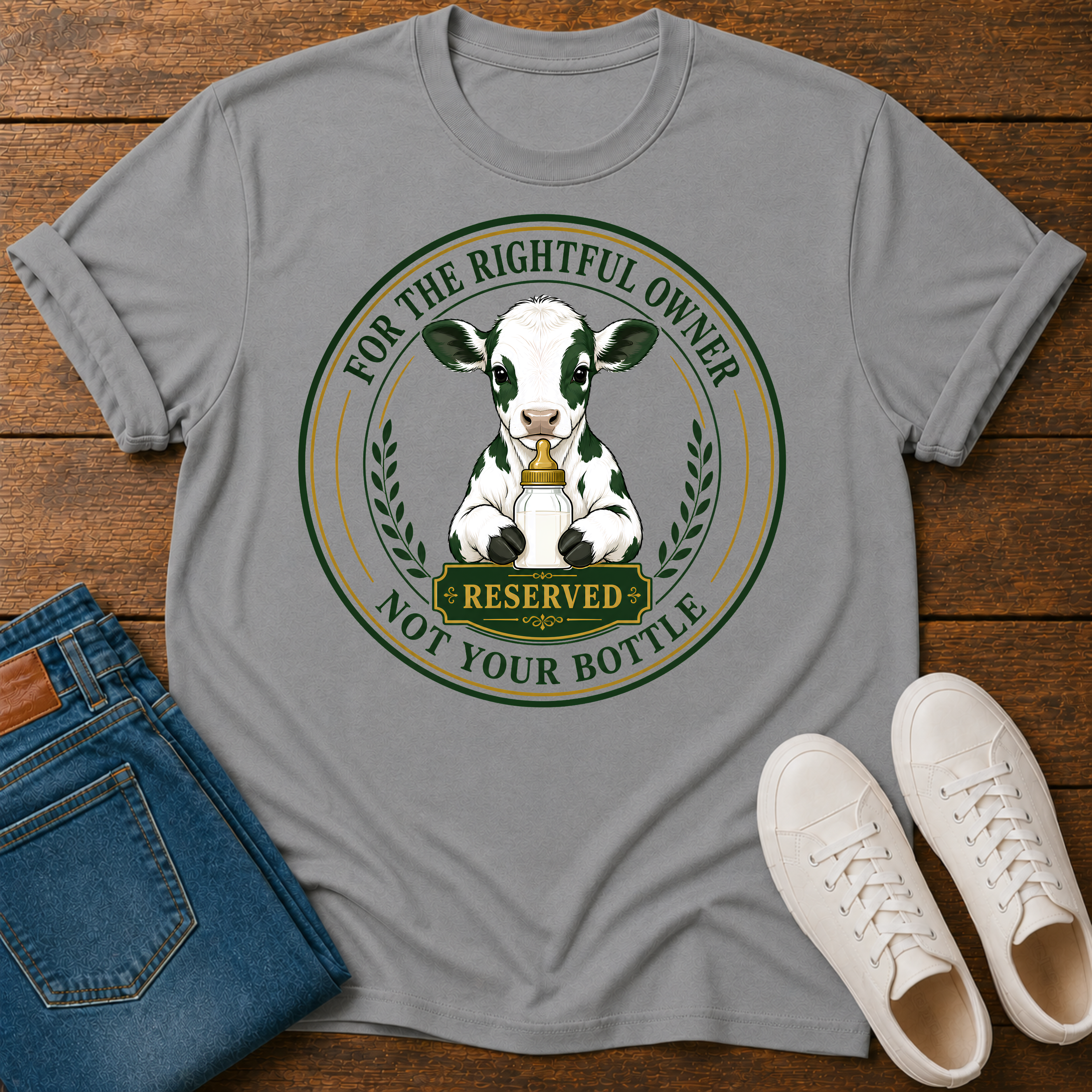 A gray short-sleeve graphic T-shirt laid flat on a wooden surface, styled with folded blue jeans and white sneakers. The shirt features a circular cow illustration with the text 'FOR THE RIGHTFUL OWNER NOT YOUR BOTTLE' and a 'RESERVED' sign.