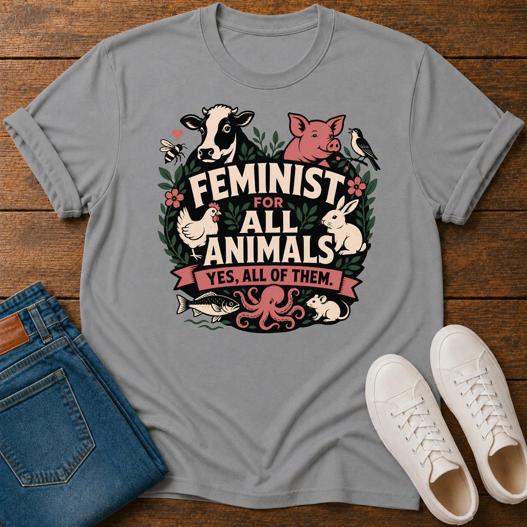 A gray short-sleeve graphic T-shirt laid flat on a wooden surface, styled with folded blue jeans and white sneakers. The shirt features a colorful front print with farm and wild animals surrounding the text 'FEMINIST FOR ALL ANIMALS YES, ALL OF THEM.