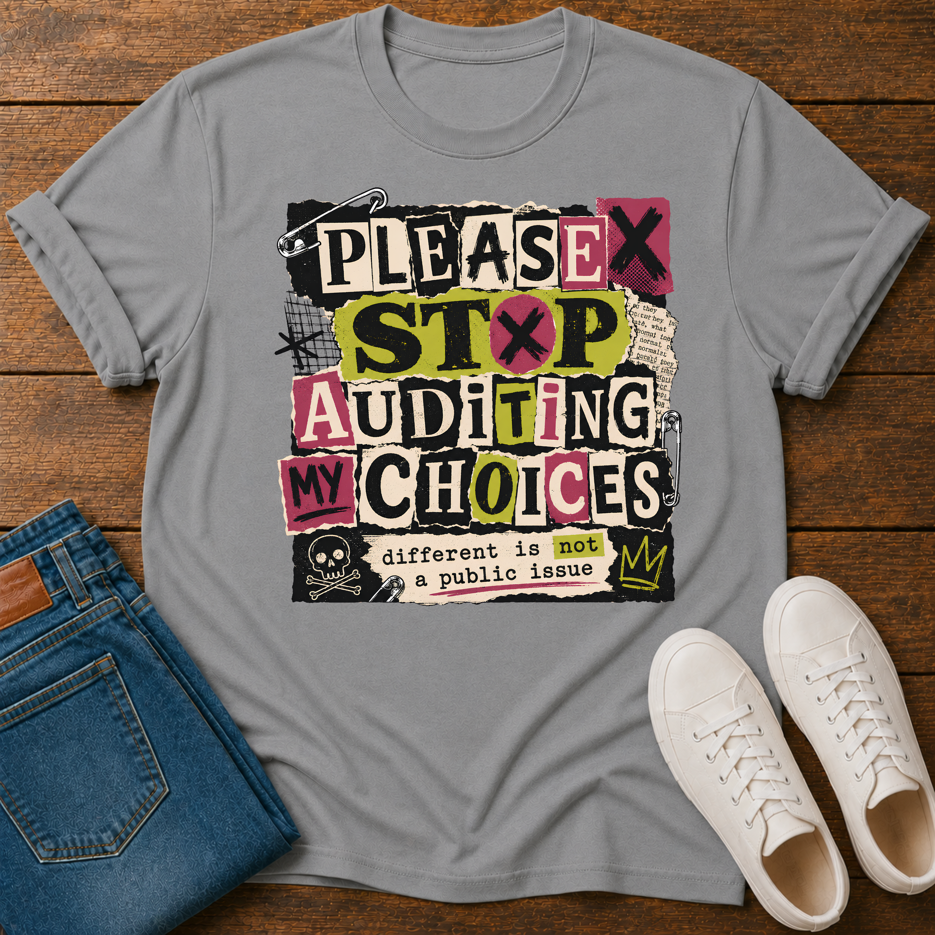 A gray graphic t-shirt laid flat on a wooden surface, styled with folded blue jeans and white sneakers. The t-shirt features a colorful, distressed graphic that reads 'PLEASE STOP AUDITING MY CHOICES' with a skull and crown detail.