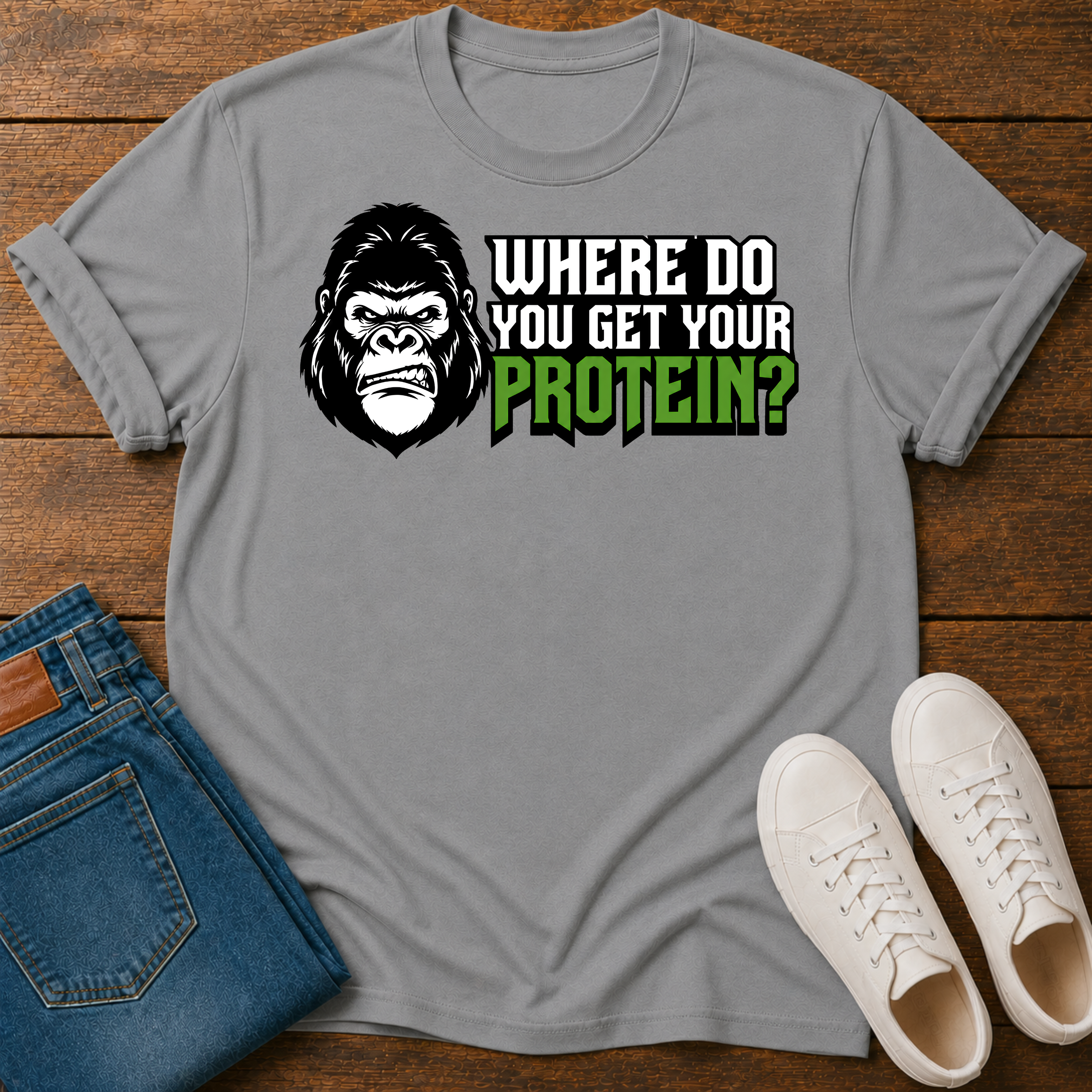 A gray short-sleeve graphic T-shirt laid flat on a wooden surface, styled with folded blue jeans and white sneakers. The shirt features a gorilla illustration and the text 'WHERE DO YOU GET YOUR PROTEIN?' in white and green.