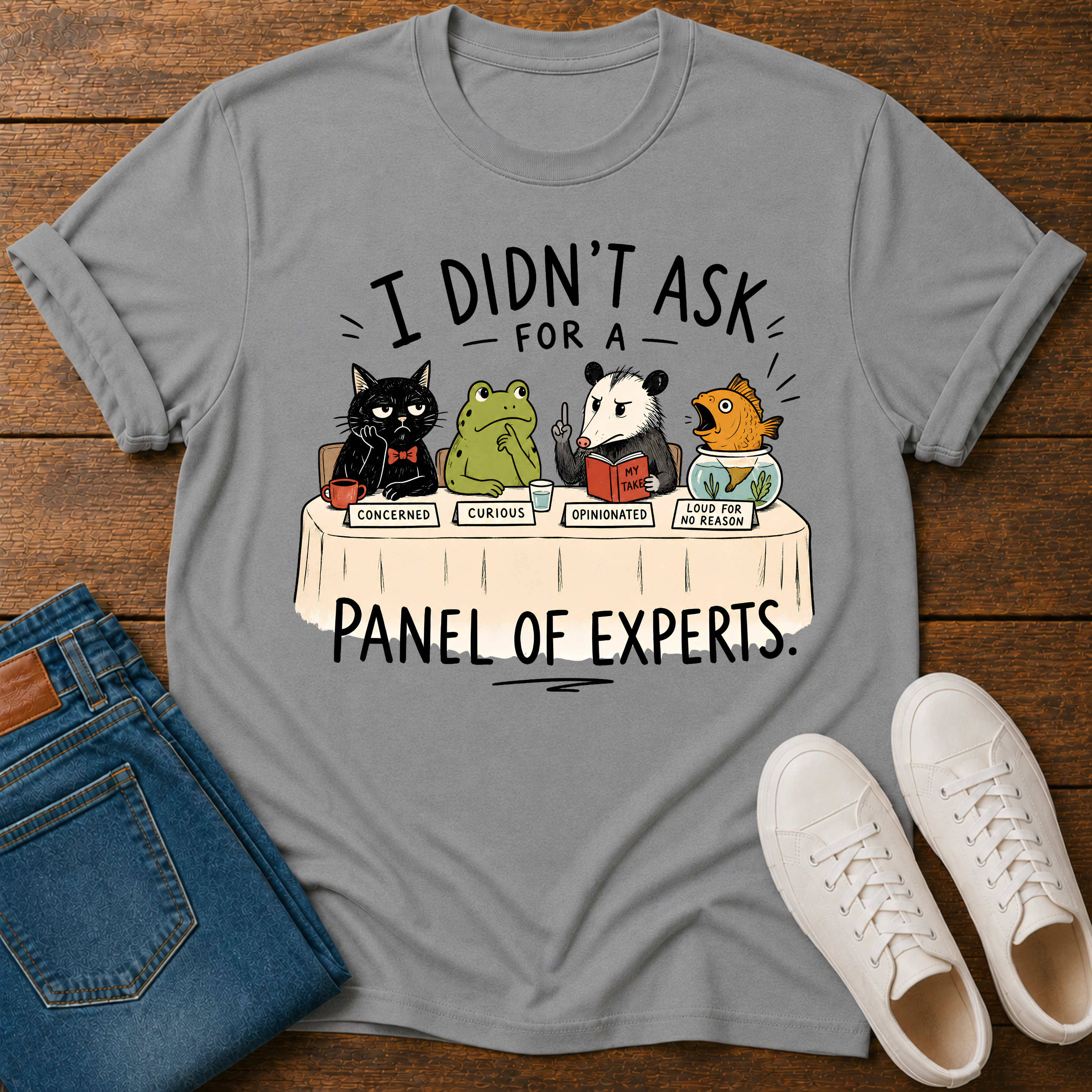 A gray graphic t-shirt laid flat on a wooden surface, styled with folded blue jeans and white sneakers. The t-shirt features a cartoon illustration of four animals (a black cat, a green frog, a panda, and a toucan) sitting at a table labeled 'PANEL OF EXPERTS' with humorous labels for their opinions.