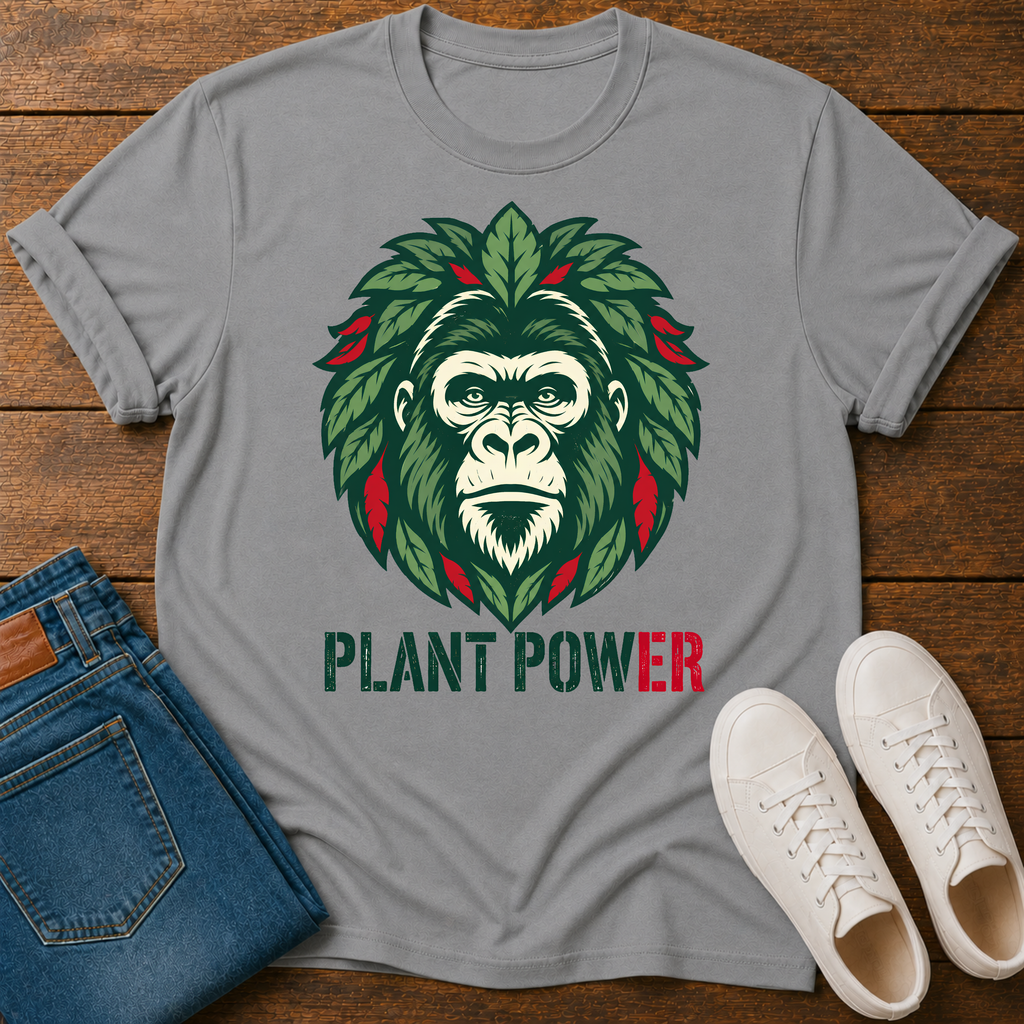 A gray short-sleeve t-shirt laid flat on a wooden surface, featuring a graphic of a gorilla face surrounded by green leaves with red accents and the text 'PLANT POWER' below. Blue jeans and white sneakers are styled around the shirt.