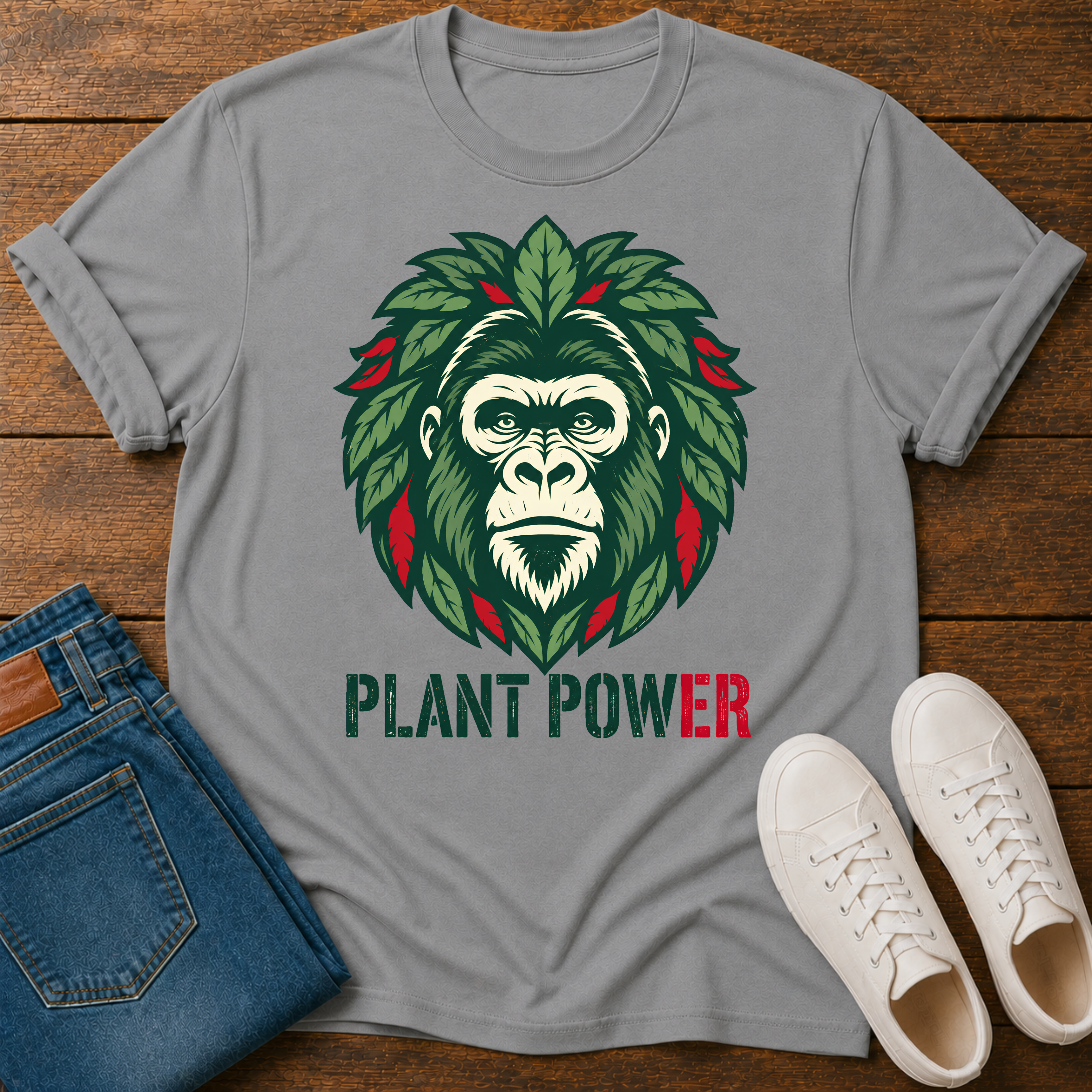 A gray short-sleeve t-shirt laid flat on a wooden surface, featuring a graphic of a gorilla face surrounded by green leaves with red accents and the text 'PLANT POWER' below. Blue jeans and white sneakers are styled around the shirt.