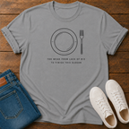 A light gray crewneck t-shirt with a black graphic of a plate and fork and the text 'TOO WEAK FROM LACK OF 812 TO FINISH THIS SLOGAN', styled in a flat lay on a wooden background with blue jeans and white sneakers.