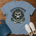 A light blue graphic t-shirt laid flat on a wooden surface, styled with folded blue jeans and white sneakers. The t-shirt features a skull graphic with a leaf pattern, crossed bones, and the text 'CERTIFIED PLANT MURDERER'.