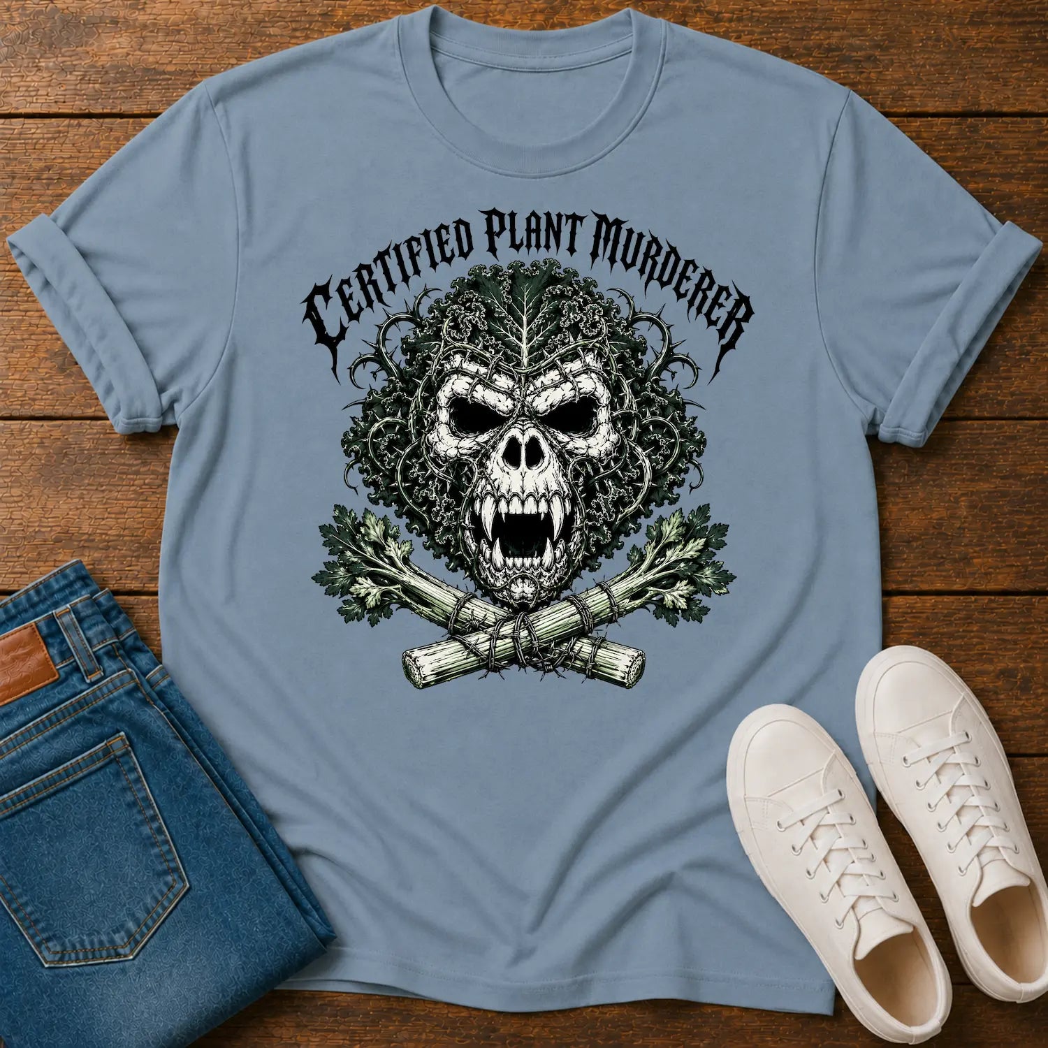 A light blue graphic t-shirt laid flat on a wooden surface, styled with folded blue jeans and white sneakers. The t-shirt features a skull graphic with a leaf pattern, crossed bones, and the text 'CERTIFIED PLANT MURDERER'.