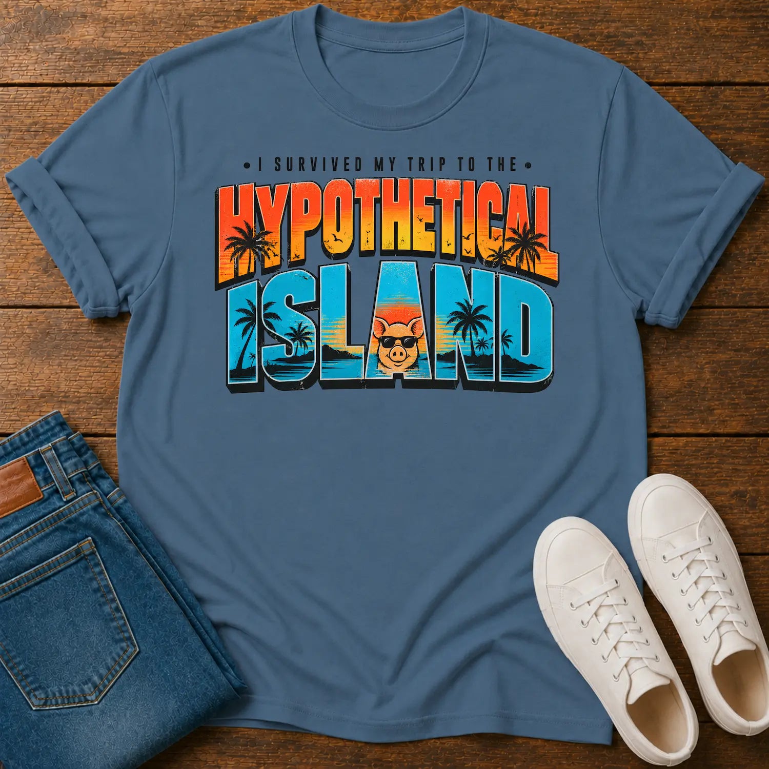 A blue graphic t-shirt laid flat on a wooden surface, styled with folded blue jeans and white sneakers. The t-shirt features a colorful retro-style graphic reading 'I SURVIVED MY TRIP TO THE HYPOTHETICAL ISLAND' with palm trees and a cartoon face.