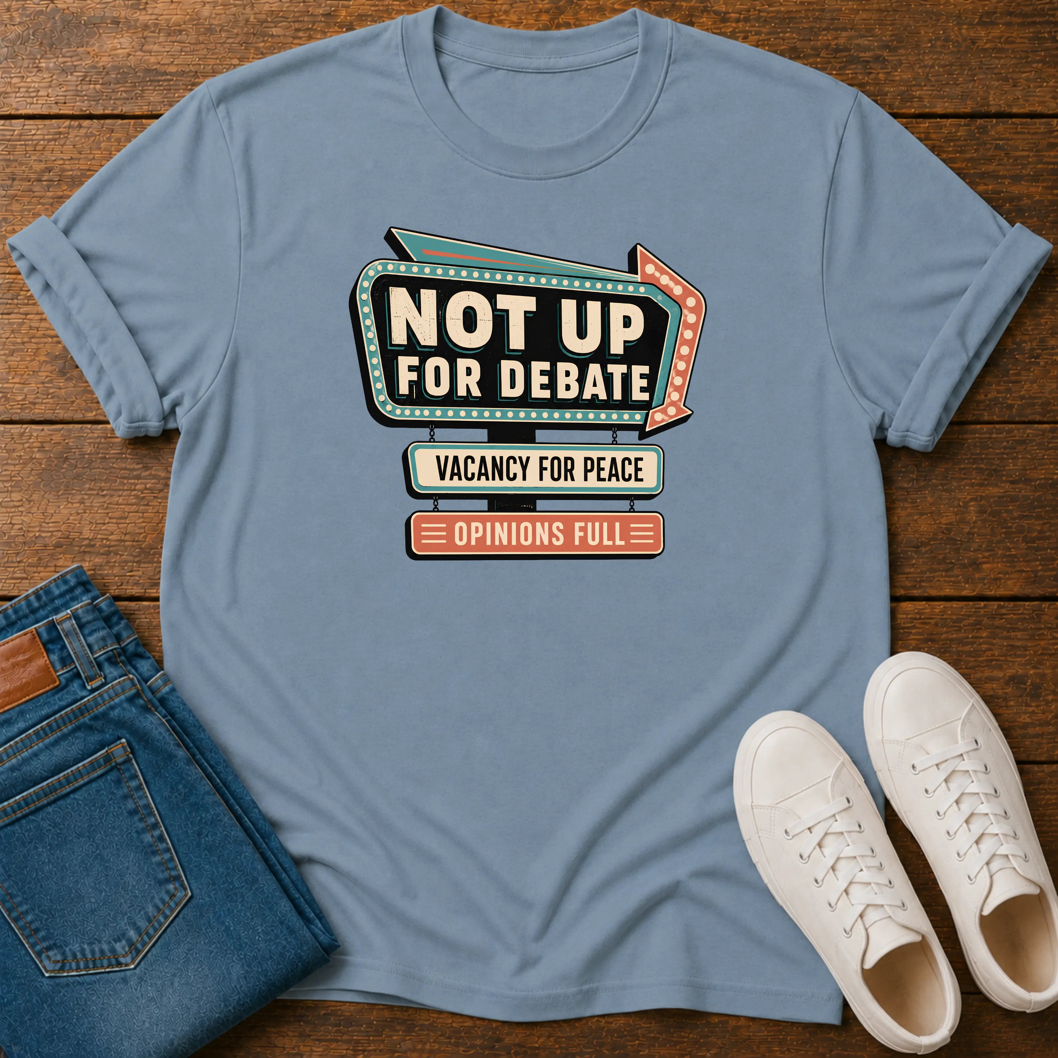 A light blue graphic t-shirt laid flat on a wooden surface, styled with folded blue jeans and white sneakers. The t-shirt features a retro marquee-style graphic that reads 'NOT UP FOR DEBATE' with 'VACANCY FOR PEACE' and 'OPINIONS FULL' underneath.