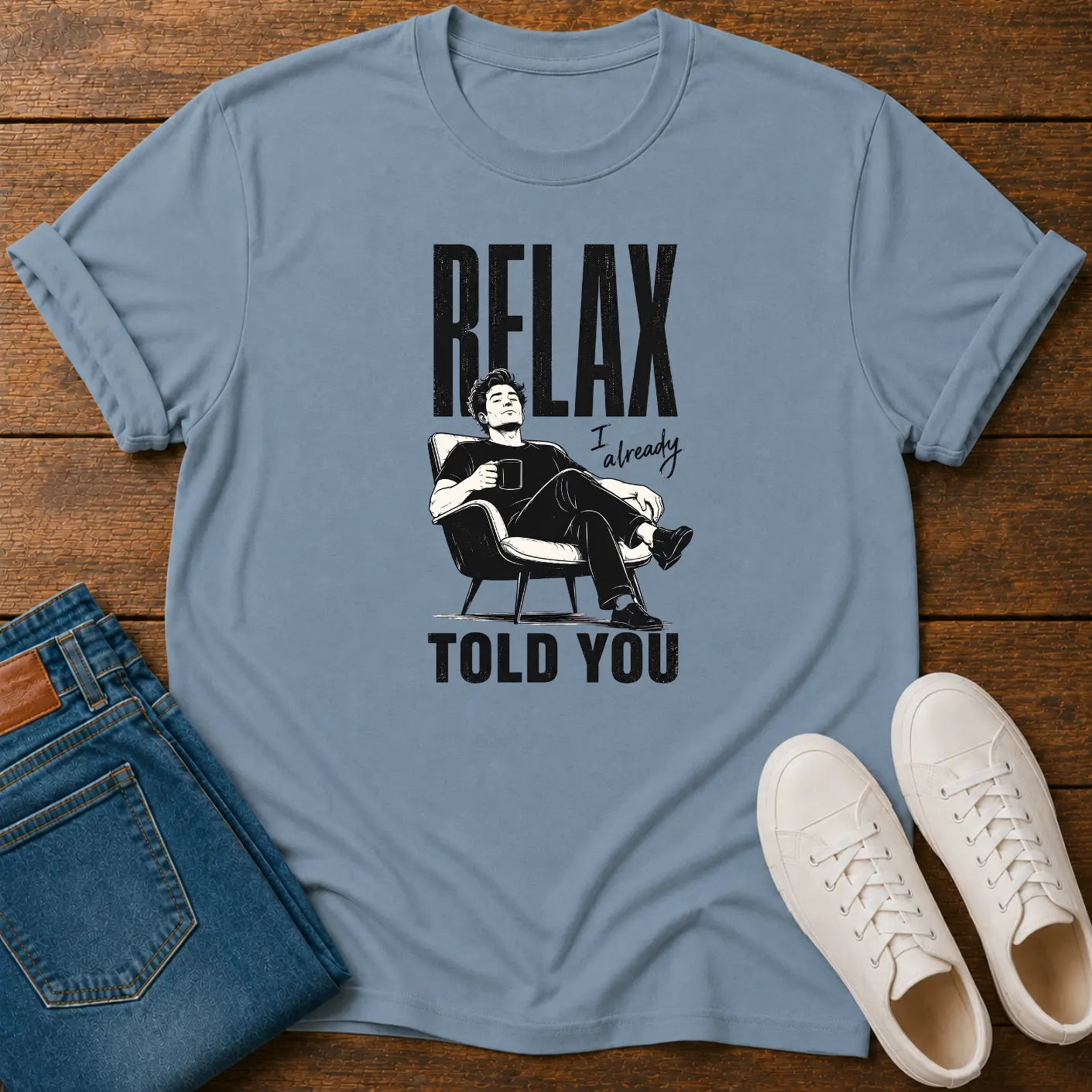 A light blue graphic t-shirt laid flat on a wooden surface, styled with folded blue jeans and white sneakers. The t-shirt features a black graphic of a man in a chair with the text 'RELAX I already TOLD YOU'.