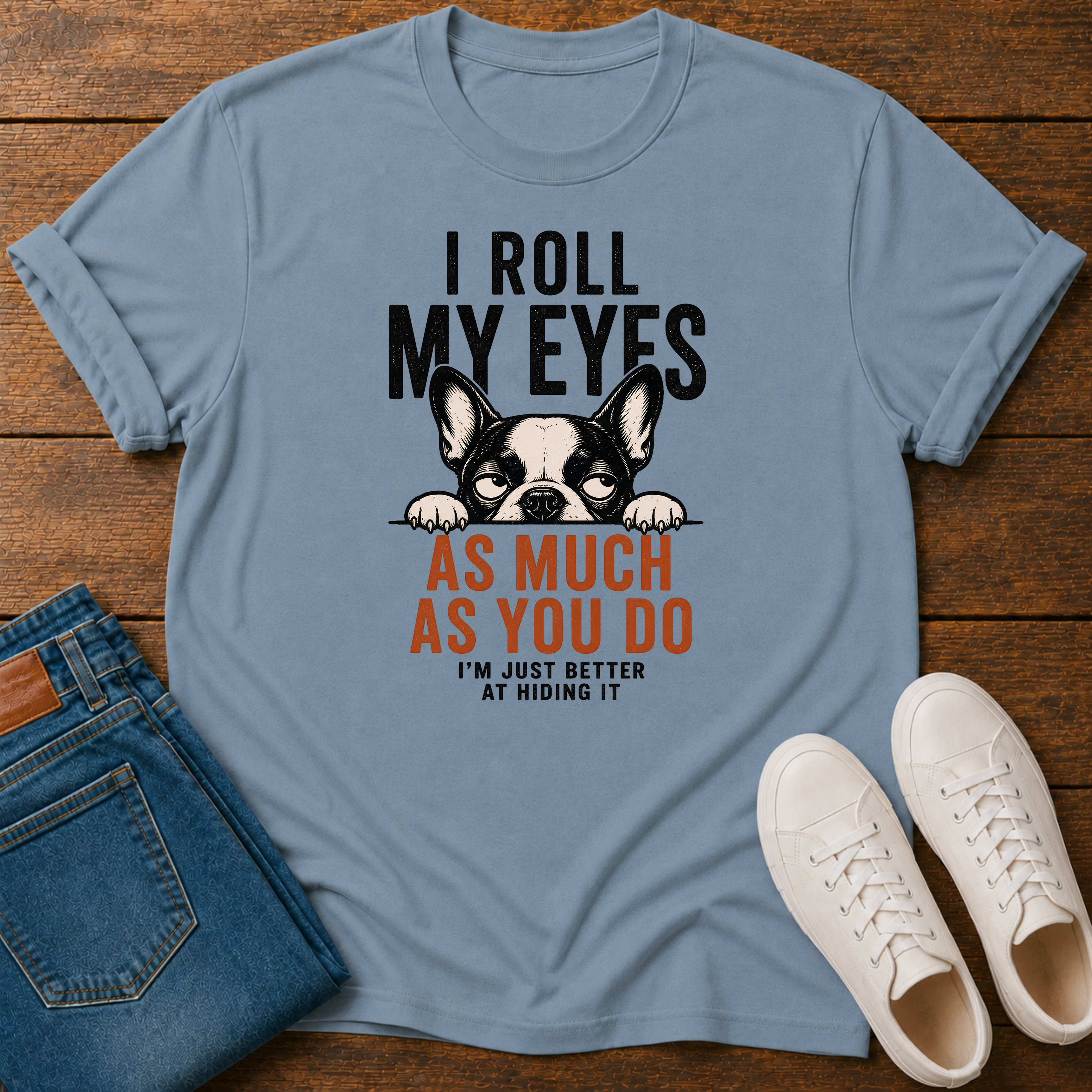 A stone blue graphic t-shirt laid flat on a wooden surface, styled with folded blue jeans and white sneakers. The t-shirt features a black and white French Bulldog graphic with the text 'I ROLL MY EYES AS MUCH AS YOU DO I'M JUST BETTER AT HIDING IT'.