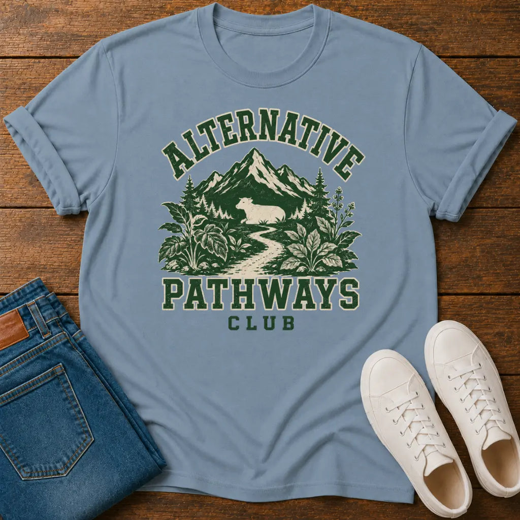 A stone blue graphic t-shirt laid flat on a wooden surface, styled with folded blue jeans and white sneakers. The t-shirt features a green and cream graphic of a mountain, forest, and a beaver on a path, with the text 'ALTERNATIVE PATHWAYS CLUB'.