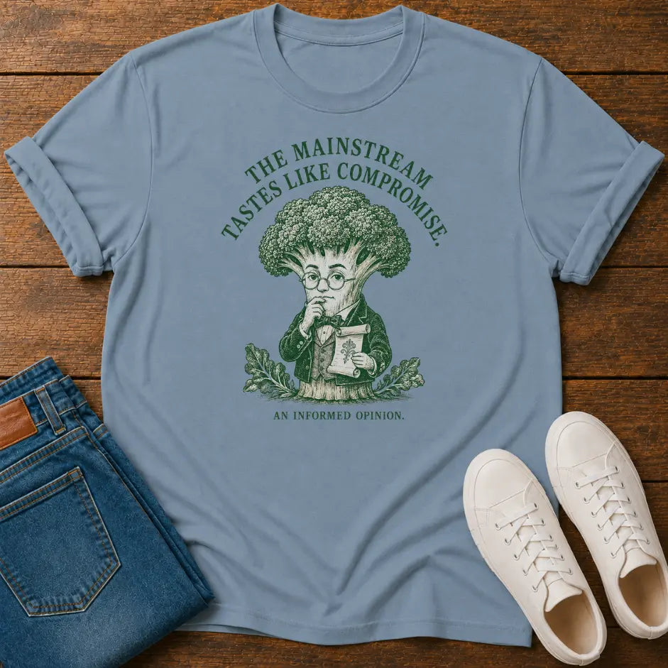 A light blue graphic t-shirt laid flat on a wooden surface, styled with folded blue jeans and white sneakers. The t-shirt features a green graphic of a broccoli character reading a newspaper, with the text 'THE MAINSTREAM TASTES LIKE COMPROMISE.