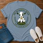 A light blue graphic t-shirt laid flat on a wooden surface, styled with folded blue jeans and white sneakers. The t-shirt features a circular graphic of a black-and-white cow with a gold bell and the text 'FOR THE RIGHTFUL OWNER NOT YOUR BOTTLE'.
