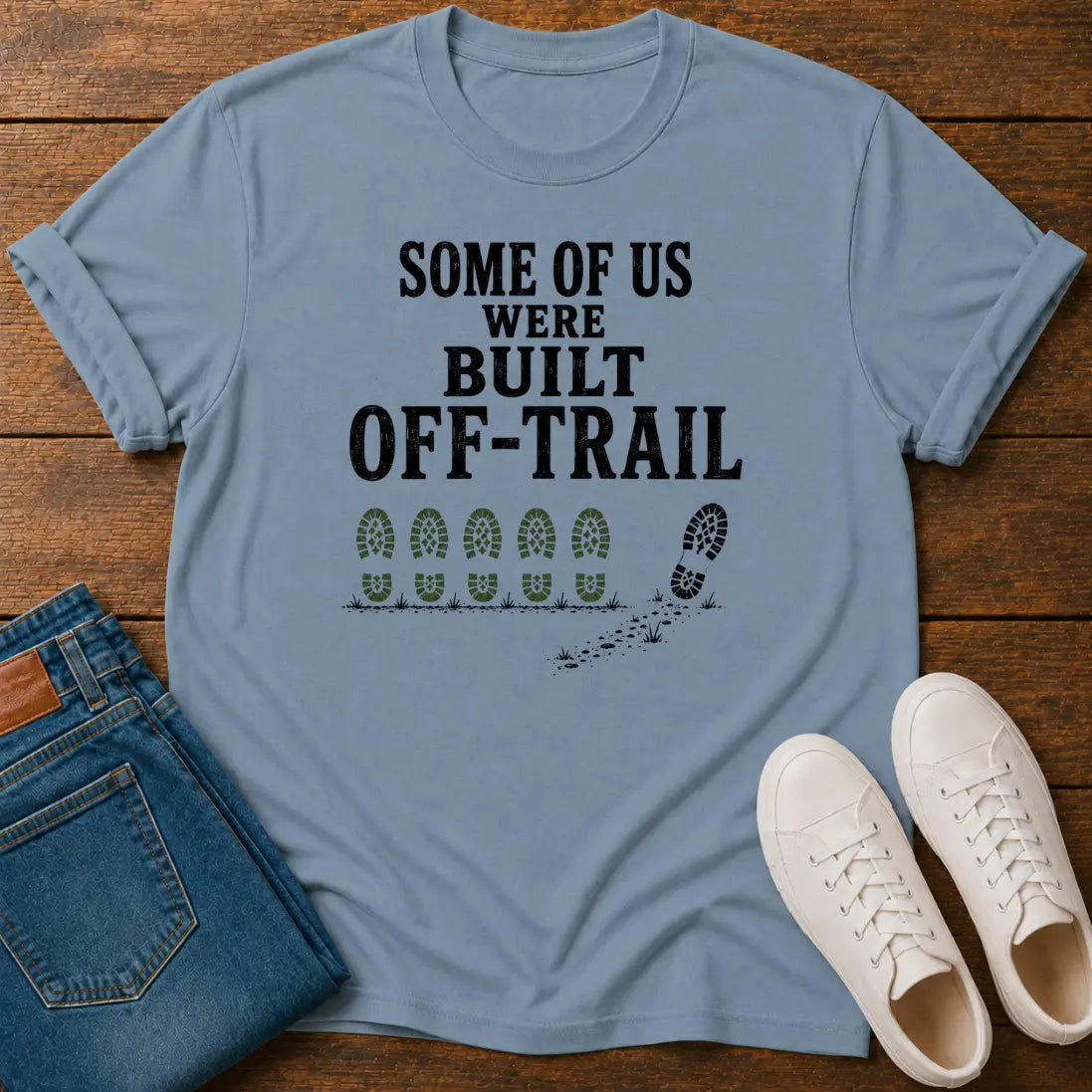 A stone blue graphic t-shirt laid flat on a wooden surface, styled with folded blue jeans and white sneakers. The t-shirt features the black text 'SOME OF US WERE BUILT OFF-TRAIL' above a trail footprint graphic.