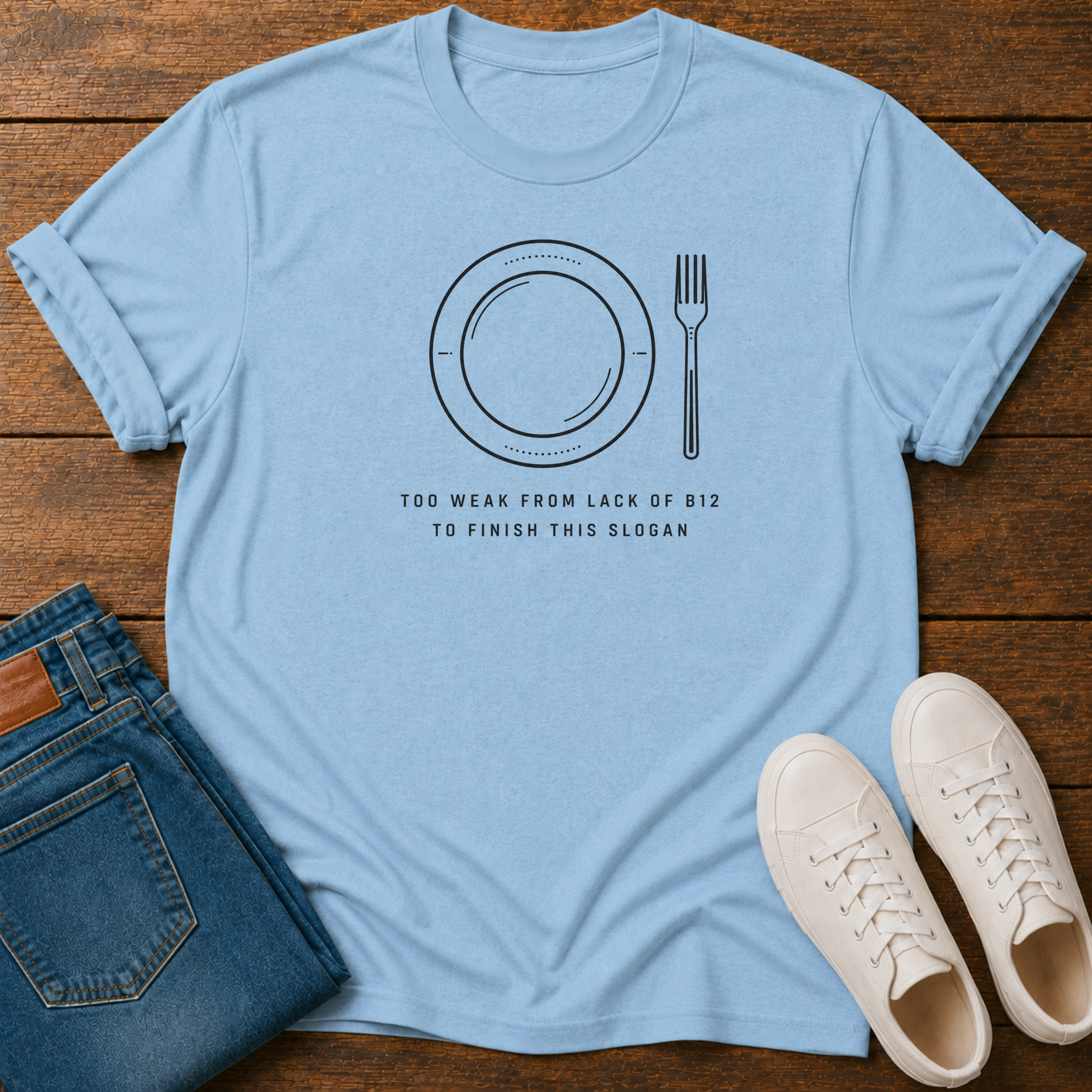 A light blue crewneck t-shirt with a black graphic of a plate and fork and the text 'TOO WEAK FROM LACK OF B12 TO FINISH THIS SLOGAN', styled in a flat lay on a wooden background with blue jeans and white sneakers.