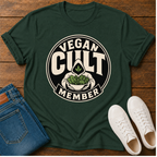 A dark green graphic t-shirt laid flat on a wooden surface, styled with folded blue jeans and white sneakers. The t-shirt features a circular graphic that reads 'VEGAN CULT MEMBER' in cream lettering, surrounding an illustration of a hand holding a bowl of leafy greens with a cannabis leaf in the center.