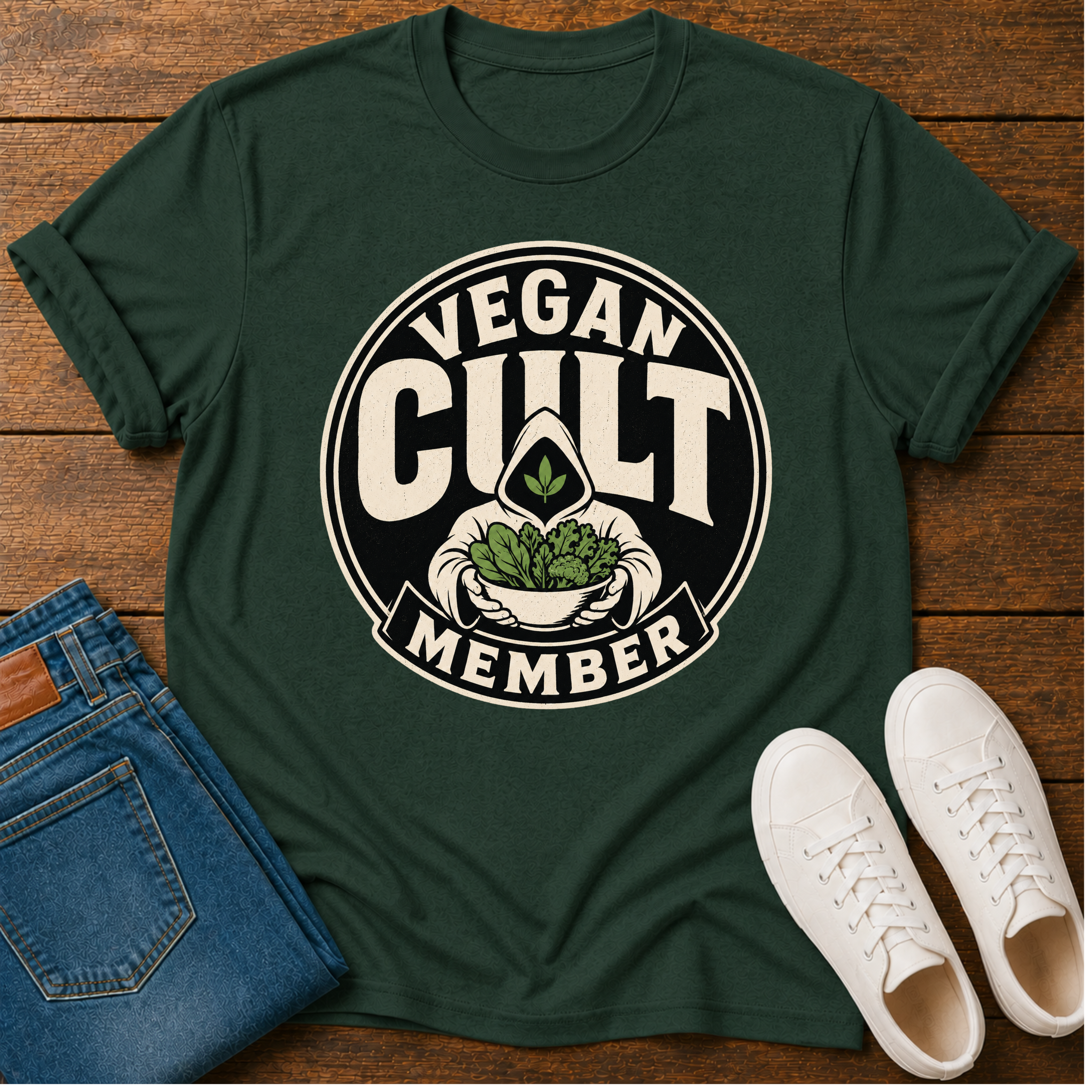 A dark green graphic t-shirt laid flat on a wooden surface, styled with folded blue jeans and white sneakers. The t-shirt features a circular graphic that reads 'VEGAN CULT MEMBER' in cream lettering, surrounding an illustration of a hand holding a bowl of leafy greens with a cannabis leaf in the center.