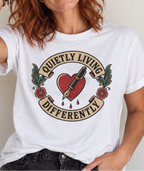 A woman with red hair wearing a white graphic t-shirt with a tattoo-style heart, pen, and banner design that reads 'QUETLY LIVING DIFFERENTLY'.