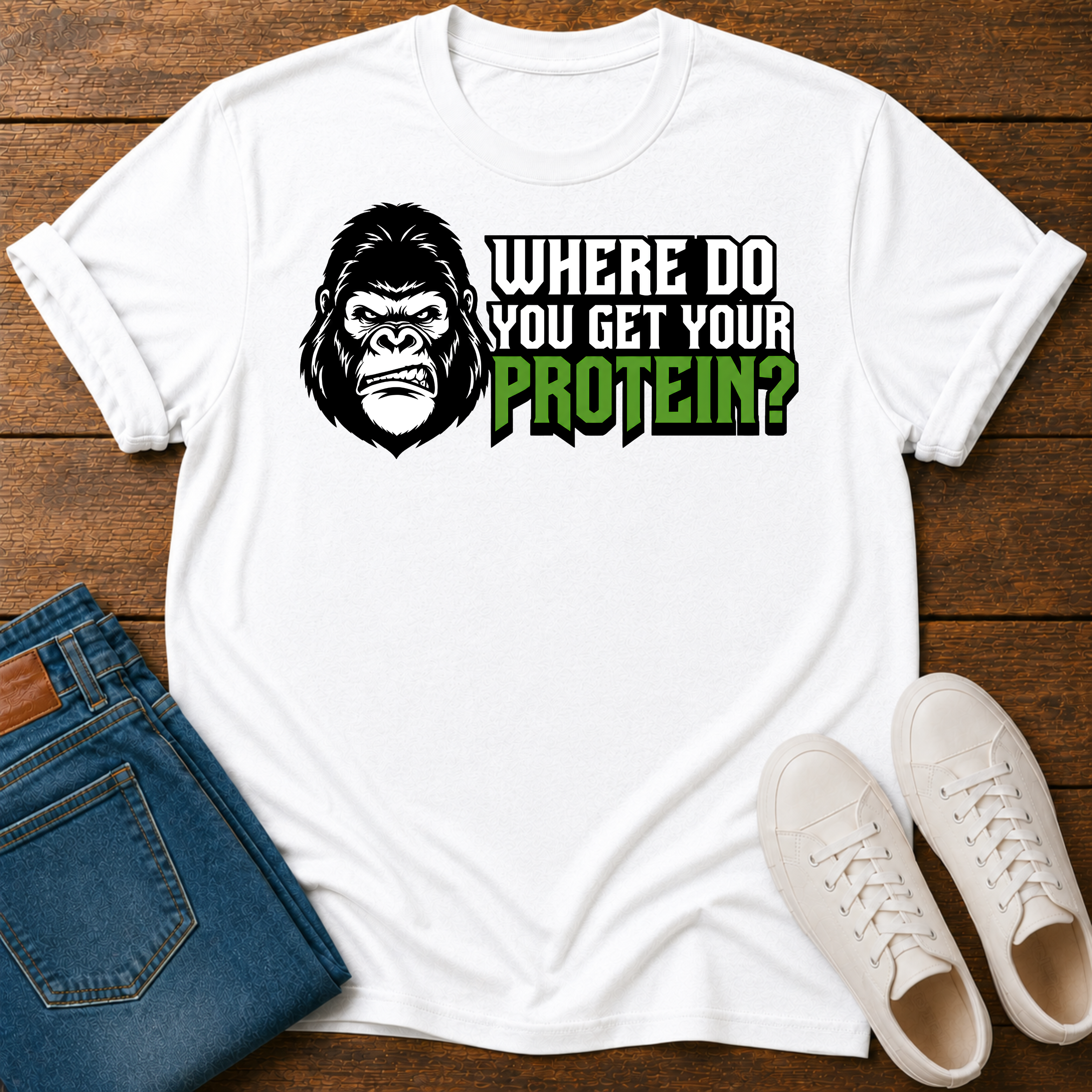 A white short-sleeve graphic T-shirt laid flat on a wooden surface, styled with folded blue jeans and white sneakers. The shirt features a large gorilla skull design with leaf details, crossed plant stems, and the text ''where do you get your protein from?"