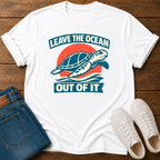 A white short-sleeve graphic T-shirt laid flat on a wooden surface, styled with folded blue jeans and white sneakers. The shirt features a retro-style turtle graphic with the text 'LEAVE THE OCEAN OUT OF IT' in blue and orange.